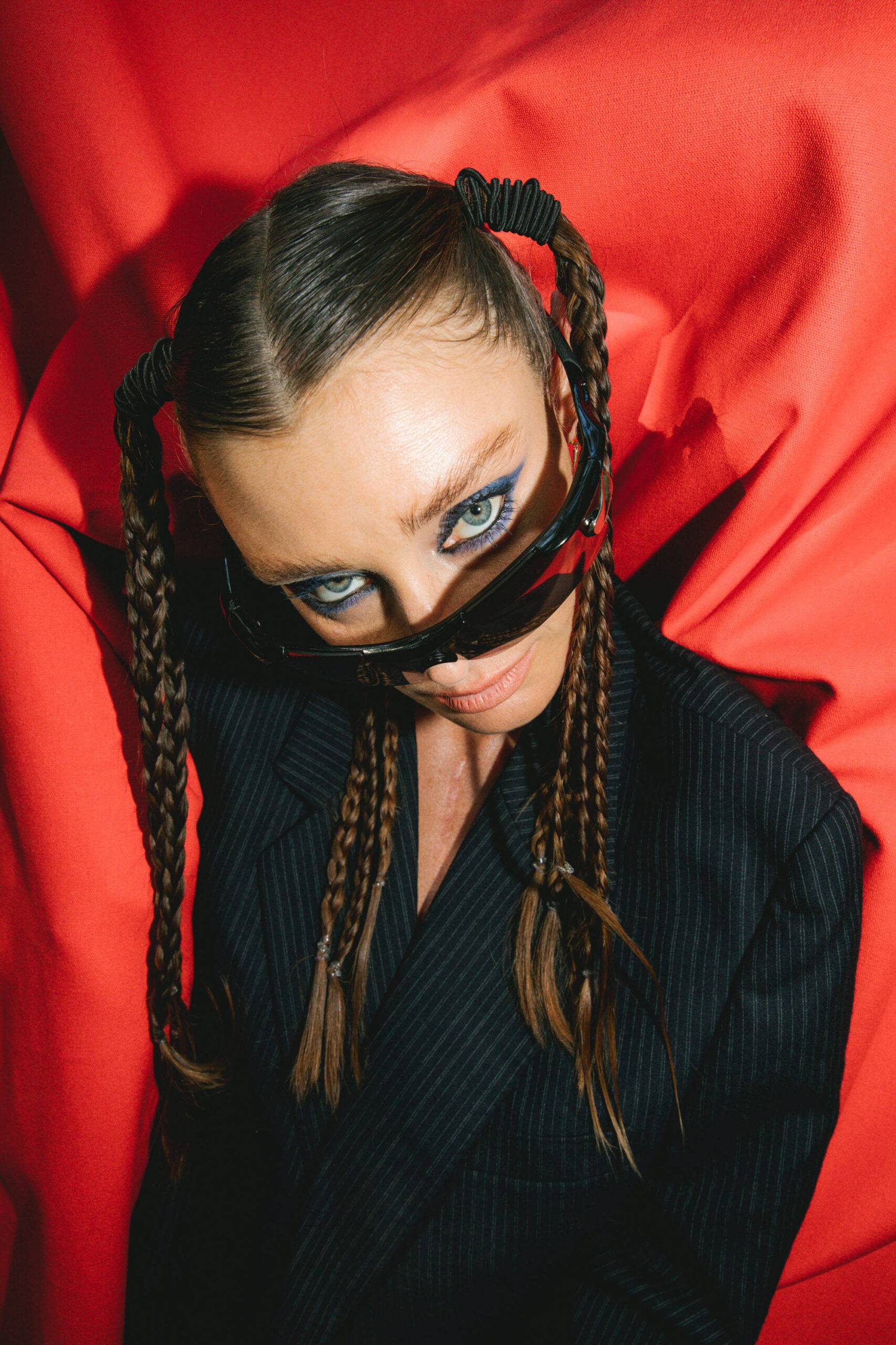 Close-up of a model with braids and black sunglasses against bright red fabric.